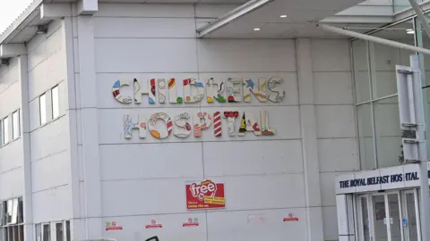 Pacemaker The exterior of the Royal Belfast Hospital for Sick Children. The outside walls are white and modern-looking and have the words 'Children's Hospital' in large colourful letters on the wall near the front entrance. The roof is green and above the doors is a section that is all glass panelling. 