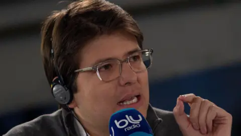 Daniel Garzon Herazo/NurPhoto via Getty Images) Miguel Uribe, wearing glasses and headphones, speaks into a microphone in 2019 while he was running for mayor of Bogota. 