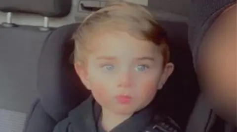 Navid Feizi A child in a car with a seatbelt on. He is wearing a black hooded top with "Nike" written on the front. 