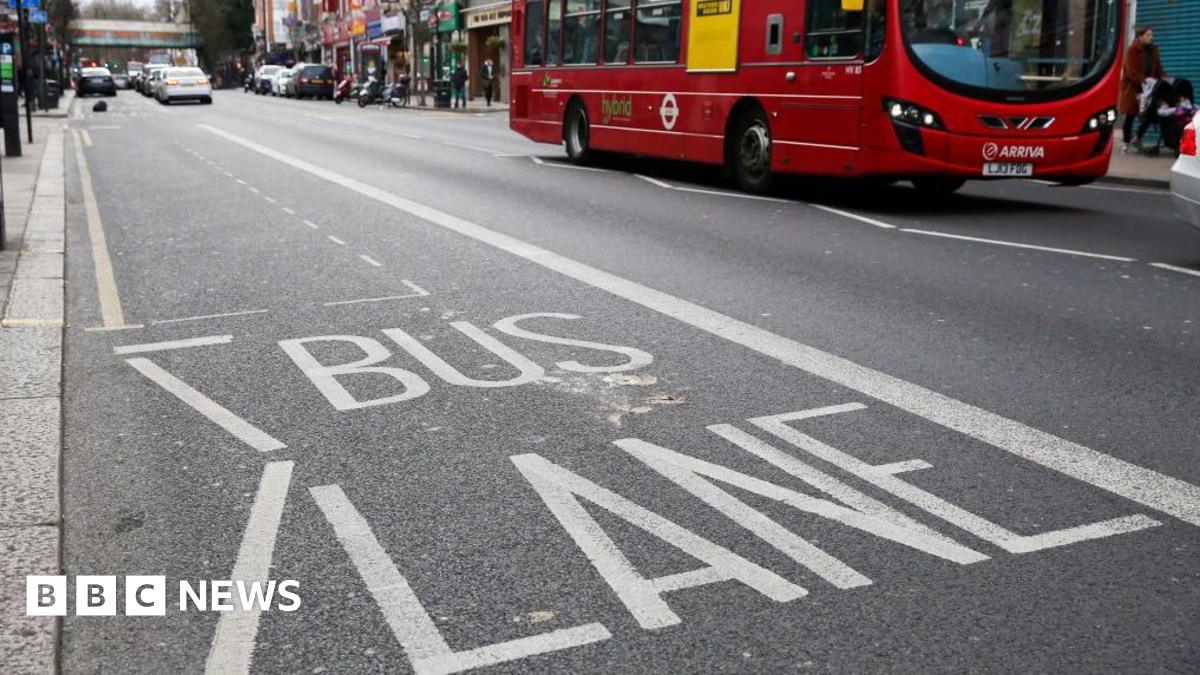 A photo of BUS LANE painted on the London road, with a red London bus in the background. There are cars on both sides of the road.