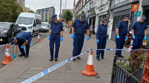 PA Media A row of six uniformed police officers search an area on Woodford Avenue in Ilford. They are in a line and looking down or to the side, working their way along a section of pavement cordoned off by police tape. Parked cars are in the road to the left and a row of businesses can be seen along the right hand side.