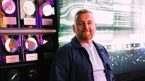 Paddy McDonnell A man with short red hair stands in front of a wall with 6 silver plaques. Over his left shoulder is a wall depicting the inside of an arena. Many people can be seen holding up phones with a flash coming out of them. The man wears a blue shirt with a grey t-shirt.