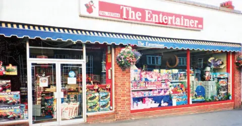 The Entertainer The first Entertainer toy shop pictured in 1981, with the window displays full of toys, hanging flowers outside, and blue and yellow awning underneath a red sign saying The Entertainer