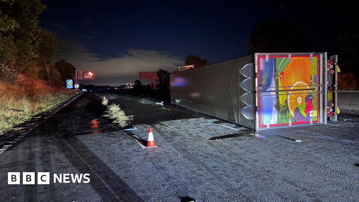 A lorry overturned on the first lane of a motorway at night. The lorry lies on its left side, illuminated by an unknown source of light. It has a silver roof and a picture of fruit on its back. An illuminated gantry in the distance is showing a closure sign. A grassy bank can be seen to the left.