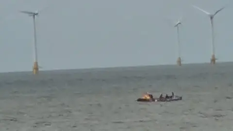 Ruper Lowe/X A picture taken by Rupert Lowe of a boat and rowing crew out at sea. In the distance there are three wind turbines. 