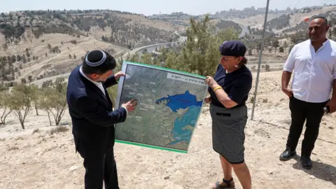 Reuters Israeli Finance Minister Bezalel Smotrich (L) helps to hold up a map showing the plans for the E1 settlement project during a visit to the area, in the occupied West Bank, on 14 August 2025