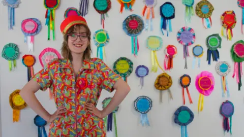 Ngaire Gape A young woman in a colourful jumpsuit wearing a red hat and glasses and with her hands on her hips, standing in front of wall covered with lots of colourful rosettes