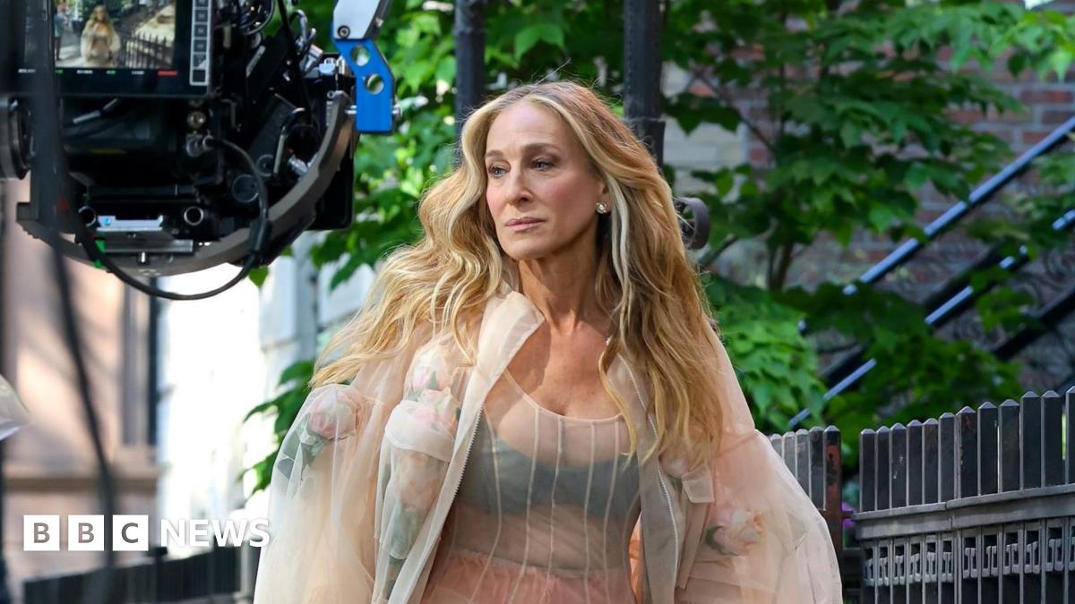 Sarah-Jessica Parker walks along a street with a large camera in front of her.