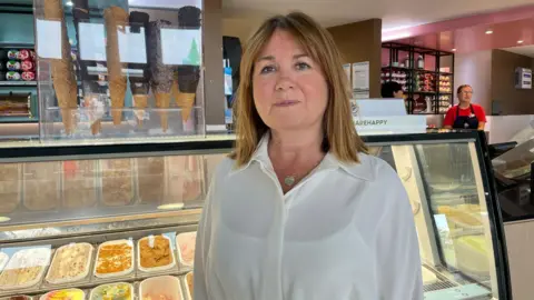Elaine Doran Jane Matthews, a white woman wearing a white blouse, stands in front of an ice cream counter. 