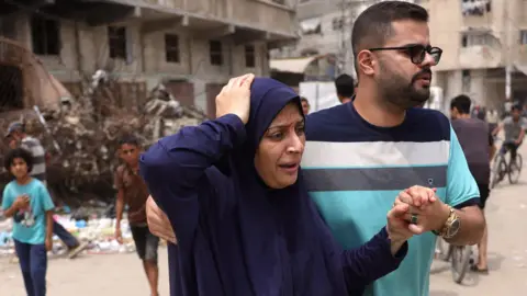 BASHAR TALEB/AFP via Getty Images An injured Palestinian woman wearing a blue robe holds her hand to the top of her head looking in pain as a man helps her by holding her left hand with his and the right hand behind her back. He is wearing a striped light blue, grey, white and navy blue shirt, has a stubble beard and is wearing glasses. Behind them there are two children and a building whose ground floors appears to have been hit