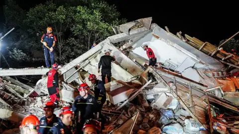 Sergen Sezgin/Anadolu via Getty Images Emergency workers in blue and red uniforms stand on a building that has crumbled from the earthquake. It is night time and the debris is light brightly by spot lights.