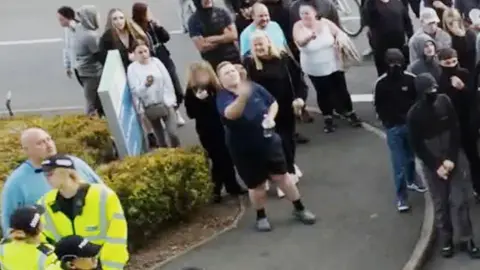 A CCTV image showing Amy Hodgkinson-Hedgecox, in dark shorts and a T-shirt, standing in a crowd of people in the Holiday Inn Express car park in Tamworth. A few police officers can be seen in front of her to her right, and to her left there are some young people who are masking their faces. Hodgkinson-Hedgecox is looking up at the hotel and gesturing with her hand, with the exact gesture having been blurred.