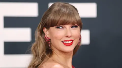 Getty Images A woman with blonde hair, red sparkly earrings and red lipstick smiles over her shoulder at camera.
