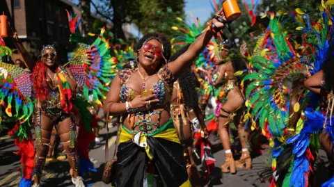 PA Media A group of women in bright costumes perform in the street. One woman at the front appears to be singing. 