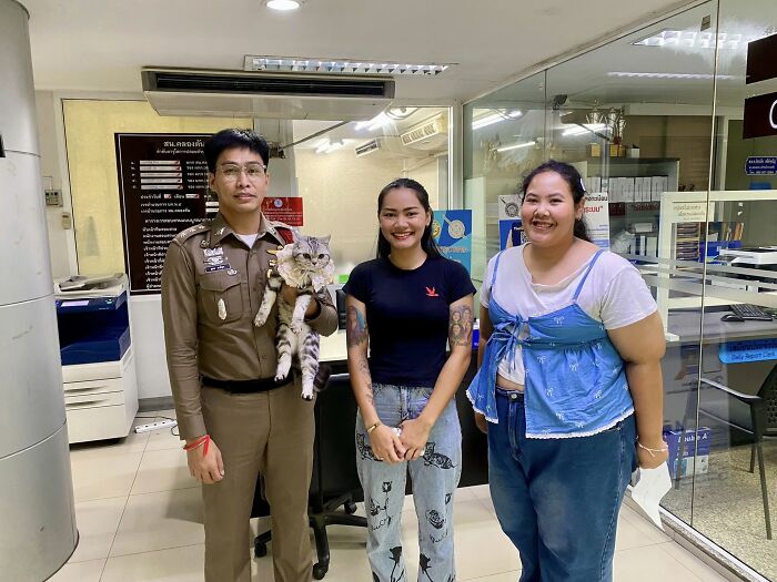 Police officer holding a cat arrested for attacking officers, standing with two smiling women inside a police station.