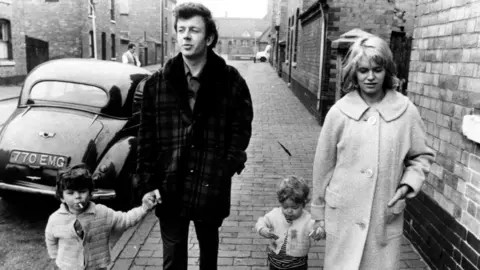 A black-and-white still image of Cathy Come Home, in which characters Reg and Cathy walk along an urban street holding the hands of two small children.