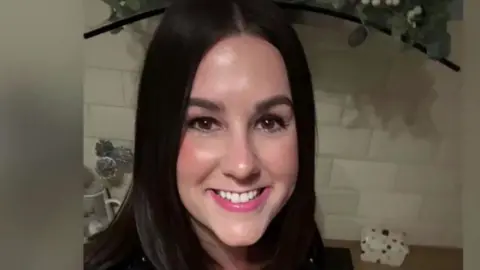 X.com A smiling Lucy Connolly looking directly down the camera. She has shoulder length, dark brown hair and brown eyes. She is seen indoors, in what appears to be a kitchen, with a white tiled splashback and work surface behind her. The edge of a hob extractor fan can also be seen behind her.