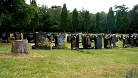 A cemetery in Tettenhall Wolverhampton. There are rows and rows of headstones marking graves and trees in the background. There is a sawn tree stump in the foreground. 