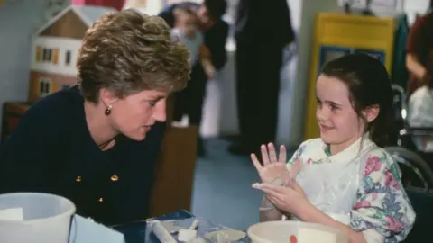 Getty Images Princess Diana, wearing a blue Chanel suit, speaks to a young girl playing with play-doh. The girl, who is looking at Diana, is wearing a floral jumper and has a sphere of play-doh in her hands.