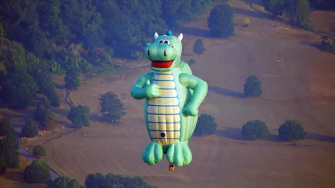PA A green dragon-shaped hot air balloon with a yellow belly hangs above Bristol in the pale morning sun. The dragon has a large red mouth smile on his face and is holding his thumb up, with his other hand on his hip. Behind him is a field, brown from the summer sun, dotted with mature trees on the edge of woodland.