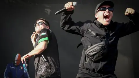 Getty Images Kneecap performing on stage. One wearing a black jacket and baseball cap. One wearing a black and green t-shirt and one wear a green, white and gold balaclava.
