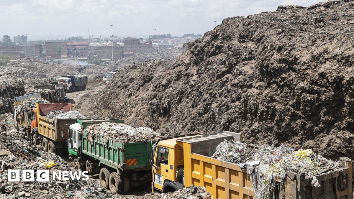 Rubbish dump trucks, green and yellow in colour, queue up laden with rubbish in a tip. To the left and right are high piles of degrading plastic and rubbish. In the background the skyline of Nairobi can be seen