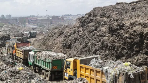 James Wakibia/Getty Images Rubbish dump trucks, green and yellow in colour, queue up laden with rubbish in a tip. To the left and right are high piles of degrading plastic and rubbish. In the background the skyline of Nairobi can be seen