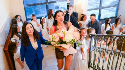 President of the Republic of Kosovo Lipa and the president walk up some stairs. They both smile as the president holds Lipa's arm. The singer holds a large floral bouquet as children in white dresses stand lining the staircase