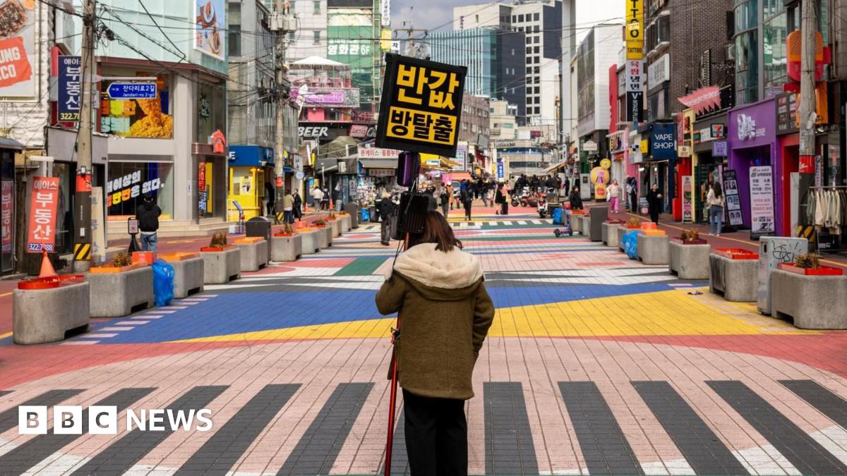 A lively and colorful pedestrian street in Seoul, filled with vibrant crosswalk designs, unique shops, and bustling activity. A woman stands in the middle of a zebra crossing in a winter jacket carrying the sign 'escape room, half price'