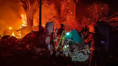 DSNS Ukraine An emergency responder stands amid debris from a strike in Ukraine, lit up by a fire in the distance.
