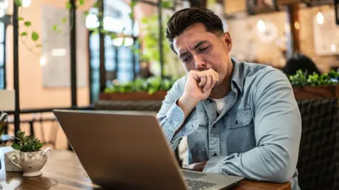 Getty Images A young man with short dark hair and wearing a denim shirt appears distracted sitting in front of a laptop, which is on a wooden table, in a large open space. In the blurred background you can make out ceiling lamps, climbing plants and other wooden tables.