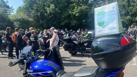 Jenny Kirk/BBC Men and women standing around in a car park, chatting. There are several parked motorbikes. A sign on a bike in the foreground says "Charity motorcycle ride 2025".