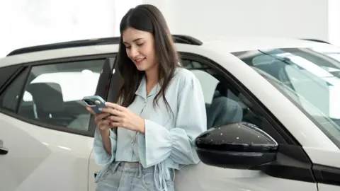 Getty Images A young woman with long straight brown hair leaning her back against a car in a showroom messaging on her mobile wearing a pale blue blouse and jeans