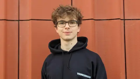 Handout Stefano pictured in front of a burnt orange textured wall. He has short brown hair, black glasses and is wearing a black hoody and smiling at the camera.