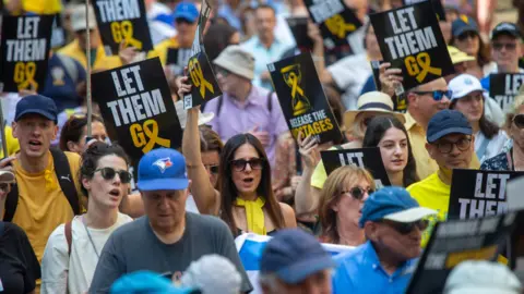 EPA A zoomed-in shot of a crowd of protesters marching through London. At the centre is a woman with long brown hair, wearing a yellow scarf and sunglasses, holding a sign that says 'let them go'. Several other protesters are holding the same sign aloft, and there are men, women and young people in the crowd.