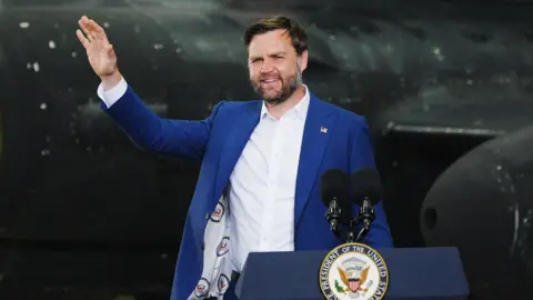 PA Media JD Vance stands waving at a lectern in front of a black US military plane in a hangar at RAF Fairford. He wears a blue suit jacket and white shirt. The lectern has two microphones and the emblem of the US Vice President's Office.