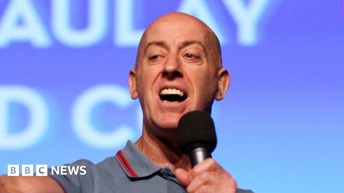 Gary Little speaks into a microphone - he is bald and wears a light blue polo shirt.