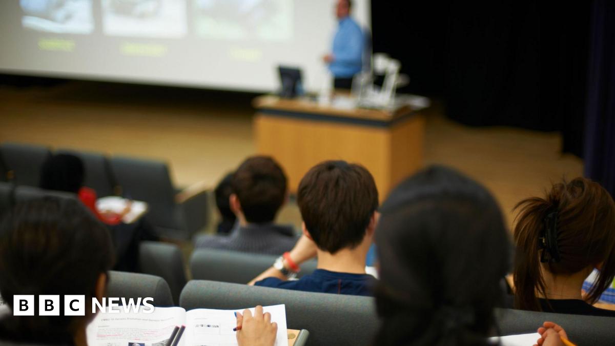 A picture taken in a university lecture hall of several rows of students, seen from behind, with their lecturer at the front who is blurred in the background of the image. The students, most of whom have dark or brown hair, appear to be writing notes as the lecturer talks.