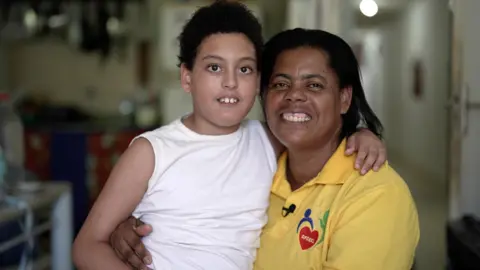 Felix Lima/BBC News Brasil Enzo and Lenice França smile at the camera. Lenice is wearing a yellow polo shirt and Enzo is wearing a white vest.