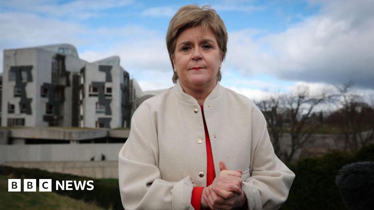 Nicola Sturgeon in white jacket and red shirt is standing rubbing her hands outside the Scottish parliament