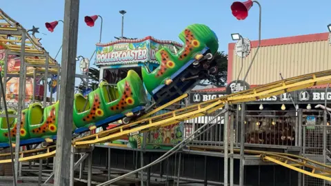 Rebecca Eccleston A side on view of a caterpillar themed rollercoaster where the front of the ride has come off the tracks. It is suspended in the air at the front as the yellow track goes out of the image. Behind there is a sign for a family rollercoaster
