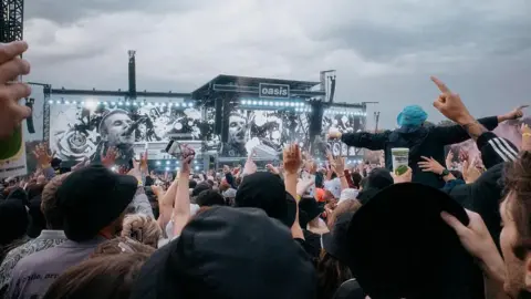 Getty Images A shot of a large crowd with their backs to the camera. The look on at a big black stage with three identical screens which record a man singing into a microphone with short brown hair. The sign in the centre of the stage reads 'Oasis.' The sky above is grey and cloudy. 