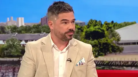 A man with dark brown hair and facial hair is being interviewed on a red sofa. He is wearing a beige suit jacket with a white shirt. In the pocket of the jacket is a handkerchief with blue and purple patterns on it