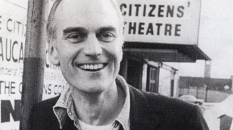Citizens Theatre A black and white shot of a man outsizxe the Citizens' Theatre in Glasgow. He is clean shaven with short hair and has a wide smile. He wears a dark blazer, a shirt unbuttoned at the neck and a light t-shirt, with a Citizens' Theatre badge on his lapel.