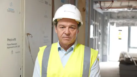 Ian Berney, a man in a white hard hat and hi-viz tabard, looks at the camera
