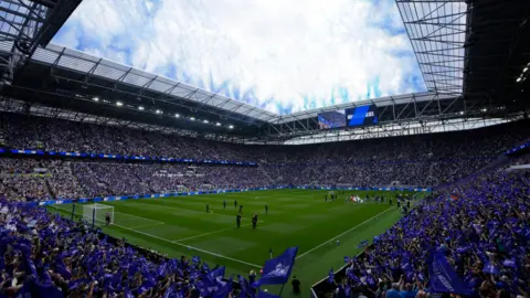 PA Media View from an interior corner of the new stadium showing fans in blue packed in all four stands around the pitch. Both teams walk out from the tunnel on the right before the match. Blue club flags are flying across both stands on left and right. Blue flares or fireworks have been let off from the roof overhead with white clouds above in the blue sky.