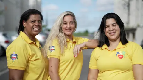 Felix Lima/BBC News Brasil Lenice França, Anne Caroline Rosa and Rute Freires stand together and smile. They are looking towards the camera. All are wearing yellow polo shirts.