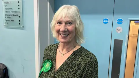 Mark Walsh/BBC Jean Glasberg smiles at the camera as she is photographed standing in a corridor with blue walls. She has short blonde hair and is wearing a green and black patterned blouse with a green rosette on her right side. She is also wearing a necklace. 