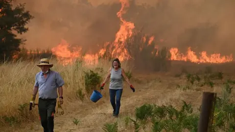 Getty Images A man and a woman try to battle a wildfire in the village of Santa Baia de Montes in the province of Ourense, northwestern Spain. They are walking away from a fierce fire in the background, and are surrounded by bone-dry grass.