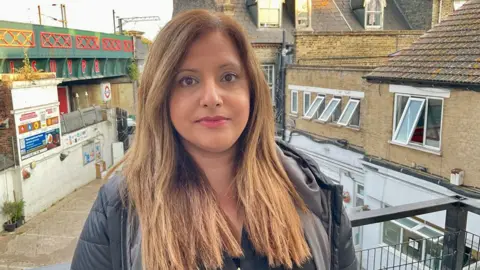 A woman in a black coat stands on a balcony in front of houses and a railway bridge in London.